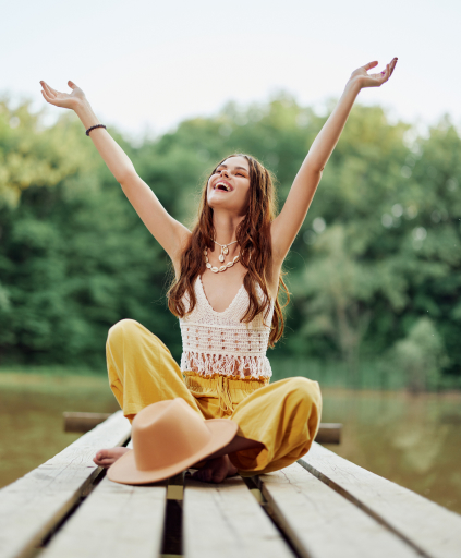 a-hippie-woman-sits-on-a-bridge-by-a-lake-with-her-hands-up-in-the-air-while-traveling-in-nature-and-smiling-in-ecoclothing-relaxed-lifestyle-1.jpg